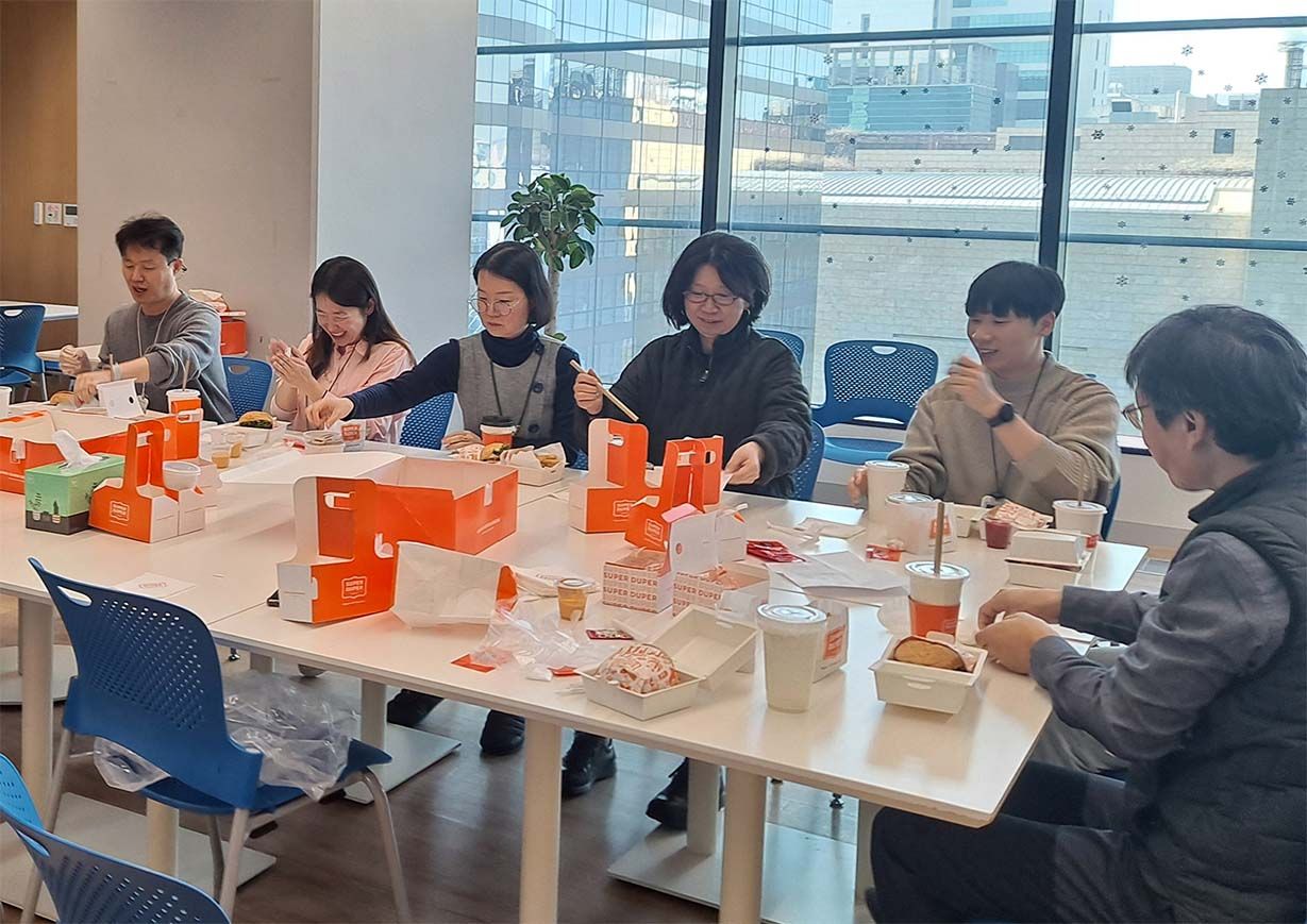 A group of colleagues enjoy a meal together at a table with takeout boxes and drinks in a bright office.