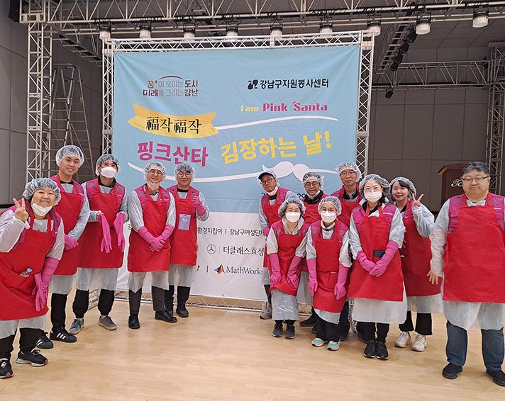 A group of volunteers in red aprons and gloves pose in front of a banner at an indoor event, smiling and making peace signs.