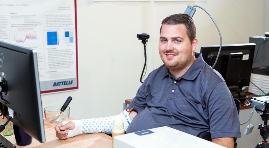 Ian Burkhart is seated in front of a computer monitor. His is wearing the sleeve on his right arm. A cable connects the implanted microelectrode array to the computer equipment. 