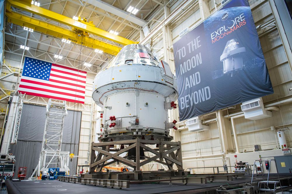 The Orion Spacecraft inside a large facility with the United States flag on one wall and a banner on another wall that says “To the Moon and Beyond.”