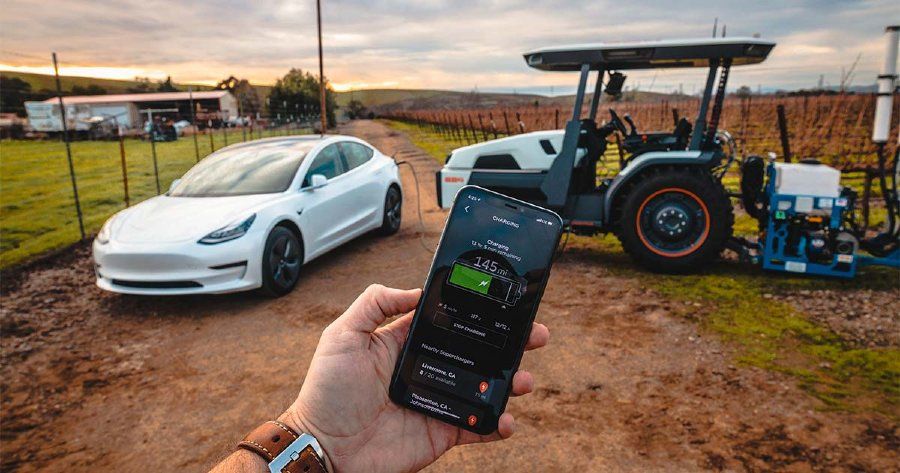 In background: Tractor is charging an electric car. In foreground: Person holding a cell phone displaying the vehicle’s charging status.